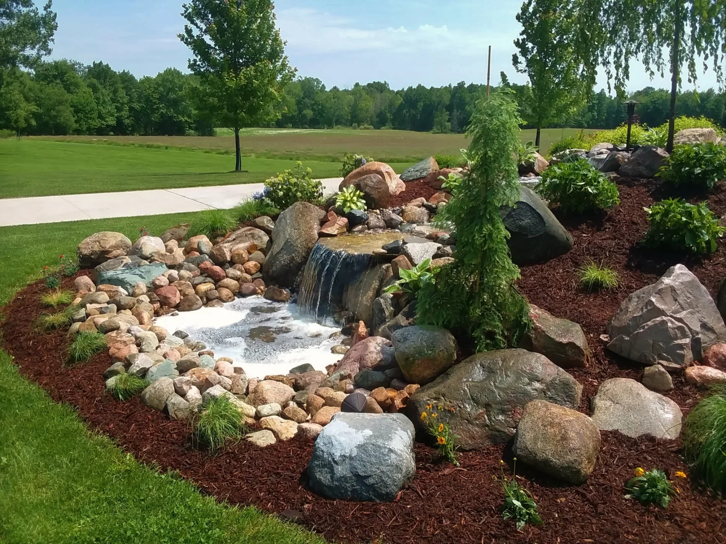 Rock-lined garden pond with a small waterfall, surrounded by mulch, stones, and green plants in a backyard landscape.