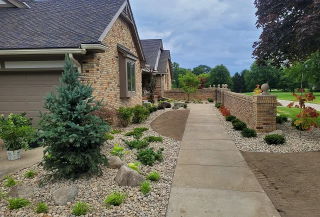 Brick house with attached garage along a landscaped front yard; a concrete sidewalk runs center, bordered by rock beds and small shrubs to the left and right.