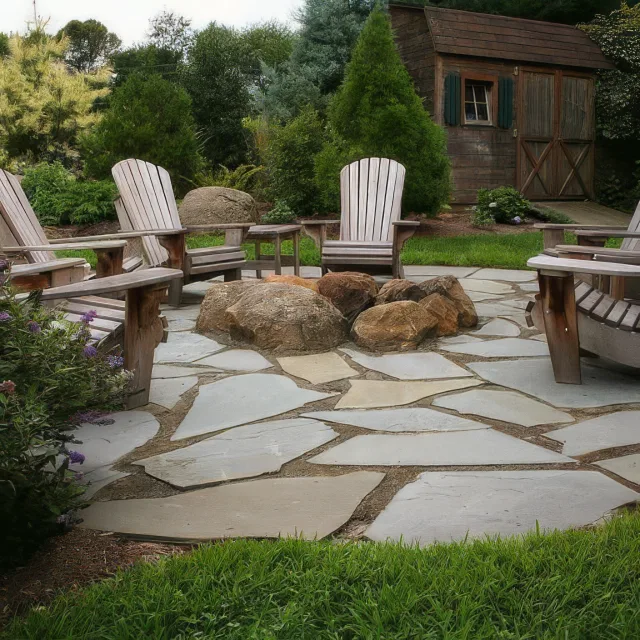 Outdoor stone patio seating area with wooden Adirondack chairs around a central rock fire pit, in a landscaped garden.